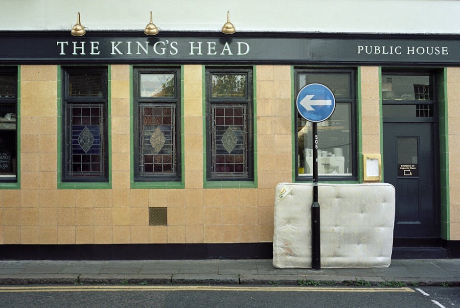 The exterior of a public house named 'The King's Head' features a beige brick facade with three vertically elongated stained glass windows, each with a decorative leaf pattern; the windows are framed in dark green. To the right, there is a dark green door with a small mail slot, and a window beside it displaying visible interior objects. A blue circular one-way traffic sign with a white arrow is mounted on a black pole situated on the pavement directly in front of the building. In front of the sign, a large, slightly dirty, indented white mattress or foam-filled padding is positioned on the pavement, leaning against the pole. The image is well-lit with natural daylight, showing a scene typical of a home relocation or furniture transport process, with the signage and exterior detailing indicating the building's nature as a public house, while the presence of the padding suggests ongoing or recent removal work. Removal Companies Twickenham is familiar with such loading or moving activities involving packaging materials, furniture, and transport preparations.