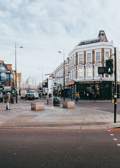A street scene in Twickenham showing a busy intersection with pedestrians walking on the pavement and crossing the road. A black and white building with large windows and decorative architectural details houses a pub named 'The World's End' on the corner. Several vehicles, including cars and a van, are waiting at the traffic lights, which display a green pedestrian symbol indicating it is safe to cross. In the foreground, cardboard boxes and a trolley are placed on the pavement, suggesting recent or ongoing moving or packing activities. The scene is illuminated by overcast daylight, and a few streetlights and road signs are visible. This image reflects a typical urban setting suitable for house removals and furniture transport, with a focus on city street logistics as part of home relocation services provided by Removal Companies Twickenham.