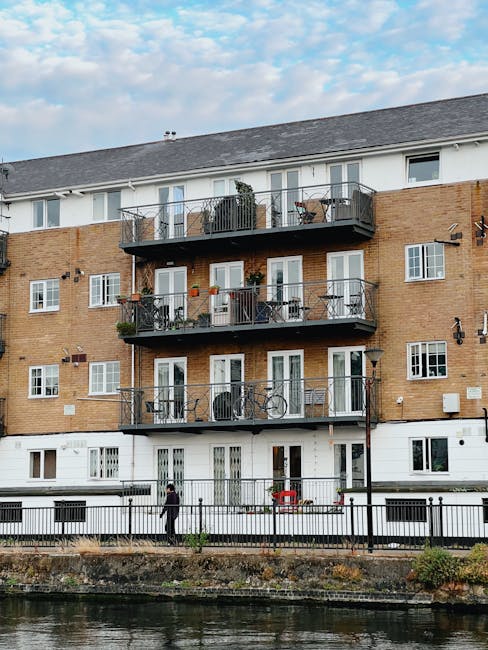 Photograph of a modern residential building situated along a waterfront, with several boats docked in the water in front. The building features multiple floors with extensive glass balconies, some wit