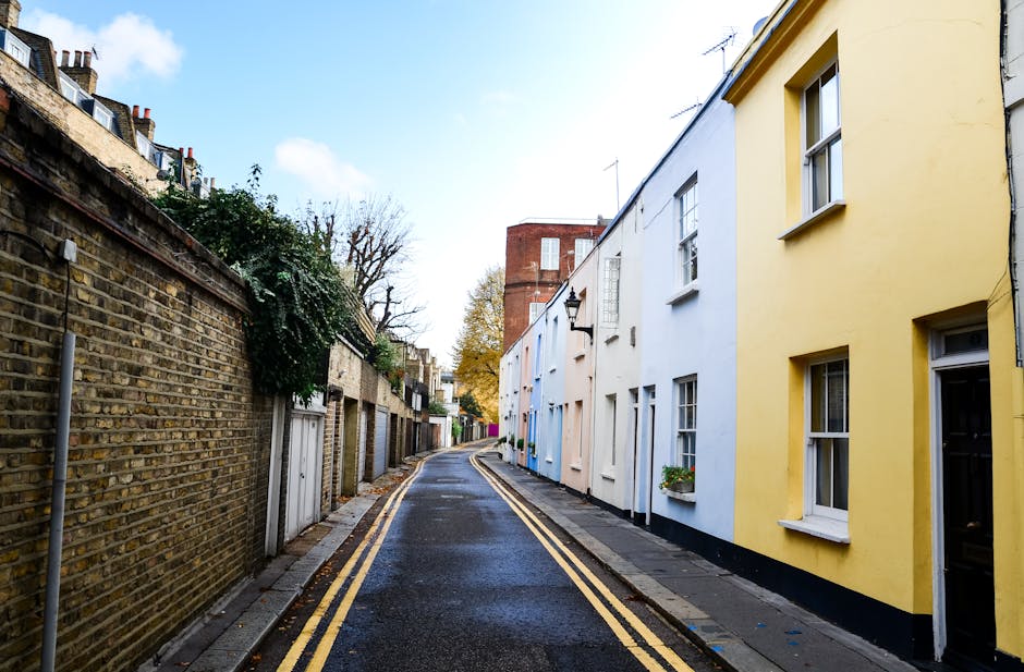 A narrow residential street in Twickenham with brick townhouses on both sides, featuring large windows and decorative facades. The street is paved with brick and has a slight incline. On the right sid