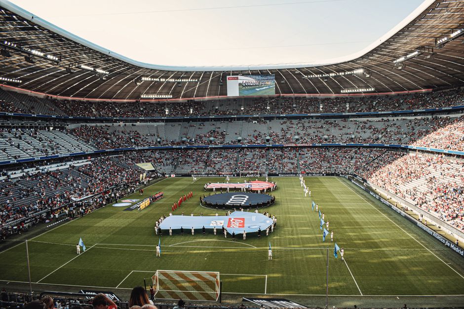 Inside a large stadium with a view of the football pitch, there are several large circular banners placed on the grass, with some featuring logos and others blank, held down by individuals wearing light-colored clothing. The seating surrounding the field is filled with spectators, and the stadium's roof structure arches overhead, allowing natural light to illuminate the scene. Along the edges of the field, there are various items such as cardboard boxes, plastic-wrapped packages, and pieces of furniture being carefully positioned or moved, likely as part of a home relocation or event setup process. Equipment like trolleys and straps are visible as movers assist with loading or unloading items. A van or removal truck is parked nearby, with its rear doors open, ready for furniture transport. The overall scene depicts a professional moving operation, possibly by Removal Companies Twickenham, preparing the stadium for a special match day event or community activity, highlighting logistics involved in large-scale removals or furniture transport within a stadium environment.